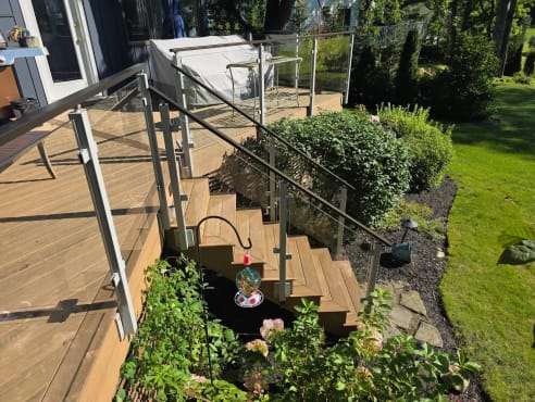 Glass panels between gray aluminum posts with cap rail line a composite deck stair into a garden.