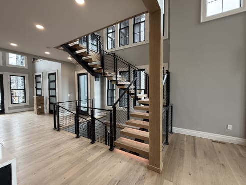 White oak treads on a black mono stringer turn at a landing with black cable railing in a bright great room.