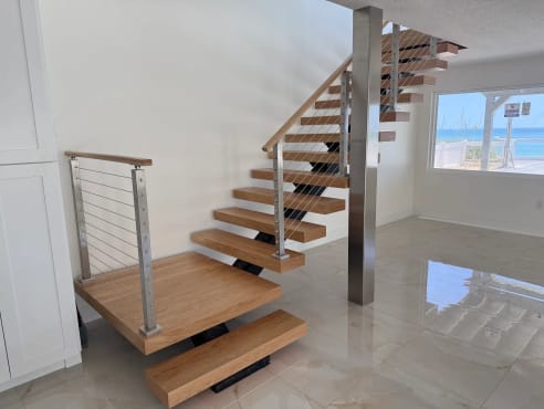 White oak floating treads on a black mono stringer start from a wide bottom landing with stainless cable railing in a bright beach home with marble floors.