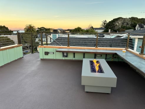 Wood-capped cable railing surrounds a rooftop terrace with a lit fire table at dusk.