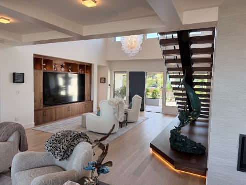 A walnut mono stair rises from a glowing plinth at the edge of an open living room with built-in media wall.
