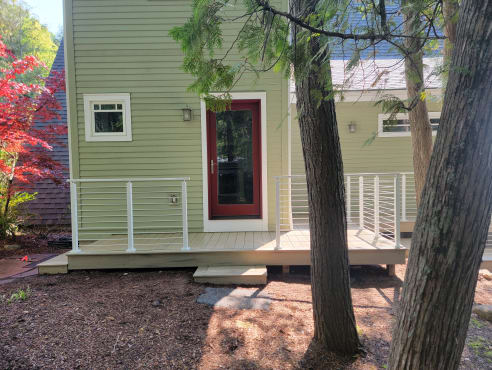 White cable railing around a small composite porch behind a green cottage among cedars.