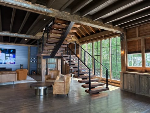 Wood floating treads on a black mono stringer switch back beneath exposed barn beams, with black cable railing and walls of windows.