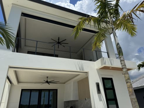 Slim gray rod railing guards the upper covered lanai of a white modern home framed by palm trees.