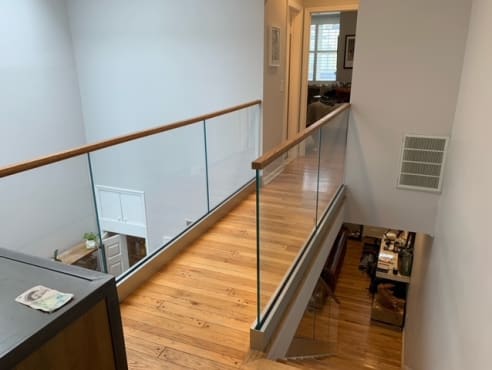 Frameless glass in a base rail with a wood cap rail runs along a pine-floored hallway above a stairwell.