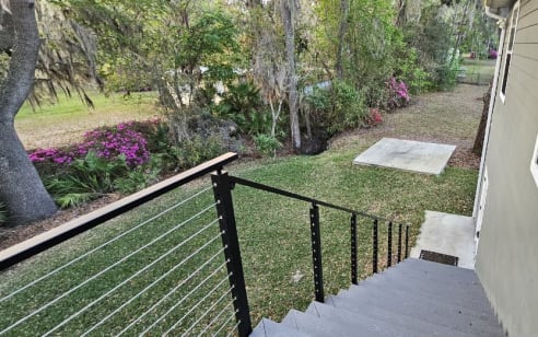 Black cable railing with a wood top rail runs down exterior steps beside a garden.