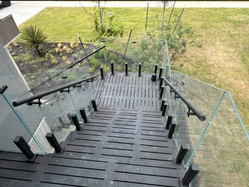 Composite deck stair from above lined with frameless glass on black talon spigots and a black round handrail.