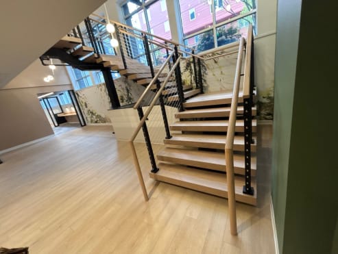 Commercial lobby oak switchback stair with black cable railing and a round light wood handrail beside tall windows.