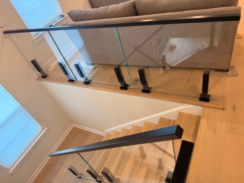 Glass panels on black talon clamps with a black handrail guard a stair opening and landing.