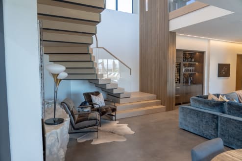 Light oak cantilevered treads rise from stacked plinth steps beside a glass railing in a modern living room.