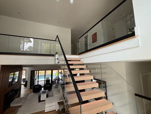 Oak floating treads with a black handrail and frameless glass rising to a glass-railed loft above a living room.