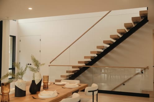 Light oak treads on a black mono stringer with a wood handrail rise along a white wall beyond a styled dining table.