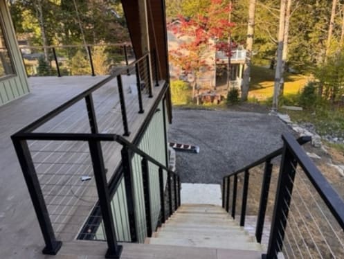 Black side-mount cable railing runs from a covered deck down an exterior stair in a wooded lakeside setting.