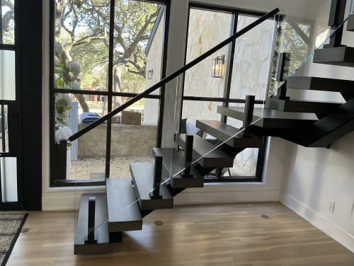 Dark-stained floating treads turn at a landing on a black mono stringer with frameless glass on talons before tall windows.