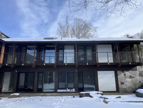 A dark-stained timber two-story home with a long upper balcony enclosed by horizontal stainless cable railing, snow on the ground.