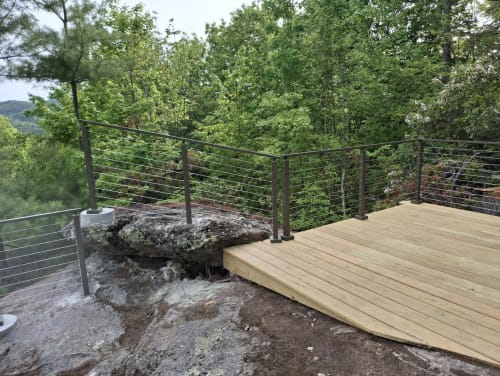 Fresh wood overlook deck on a boulder edged with dark-post cable railing above a green forest valley.