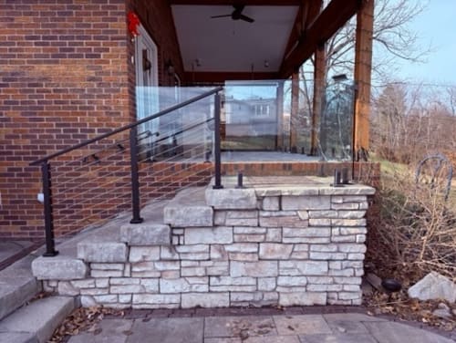 Black cable railing follows stacked limestone steps up to a brick home's covered porch.