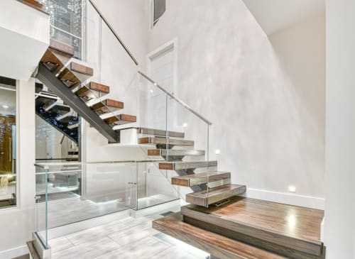 Wood floating treads on a dark mono stringer descend to a wide landing tread beside frameless glass with a wood handrail.