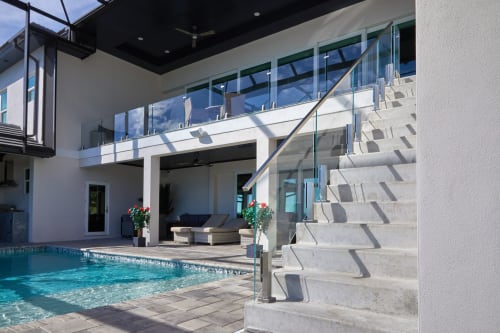 Frameless glass railings line an upper deck and exterior stair above a backyard pool under a screened enclosure.
