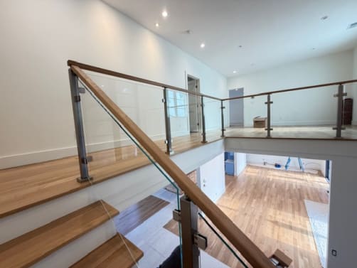 Brushed stainless posts with glass clamps and an oak handrail run up a stair and around an open balcony overlooking an oak-floored room.