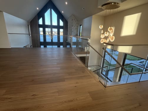 A frameless glass guard with wood cap edges an upper loft facing tall gable windows over a lake, with a globe pendant chandelier.
