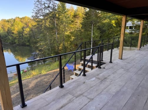 Black cable railing lines a covered composite deck stepping down toward a wooded lake.