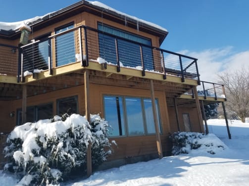 Black side-mounted cable railing on an elevated deck of a cedar home surrounded by fresh snow.