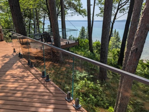 Glass panels on spigots with a top cap rail run along a wood deck high among pines above a lake.