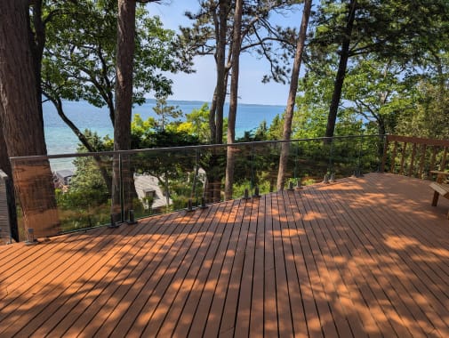 Sunlit wood deck edged by frameless glass looking through pines toward a lake.