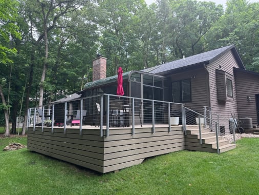 A tan composite deck wrapped in silver cable railing behind a brown-sided home in the woods.