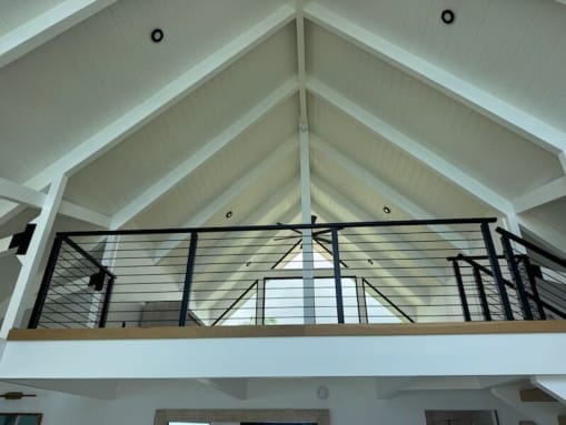 Black rod railing caps a loft balcony beneath a white vaulted ceiling with exposed rafters.