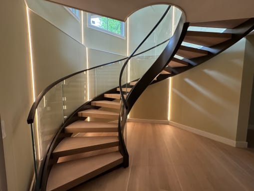 Sweeping curved stair with wood treads and dark stringers, glass railing and a curved dark handrail, lit by vertical wall LED strips.
