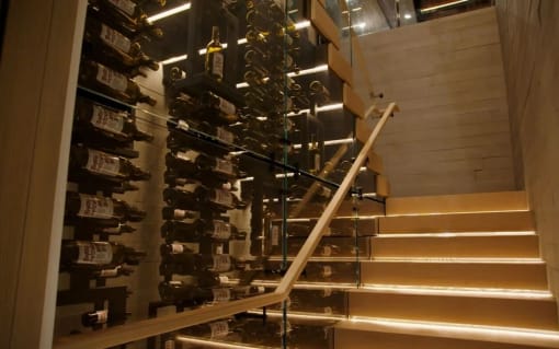 LED-lit light wood stack stair with glass railing and wood handrail running beside a glass-walled wine cellar.