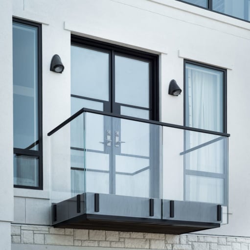 Frameless glass Juliet balcony with black top cap and fascia clamps outside double doors on a white facade.