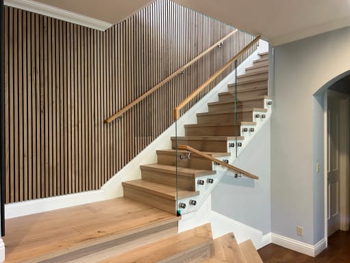 Standoff-pin mounted glass with a wood handrail follows an oak stair beside a dark slatted accent wall.