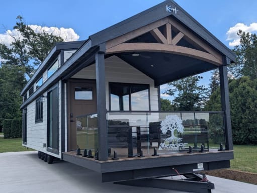Dark-sided park-model tiny home with a covered front porch enclosed by frameless glass on black spigots.