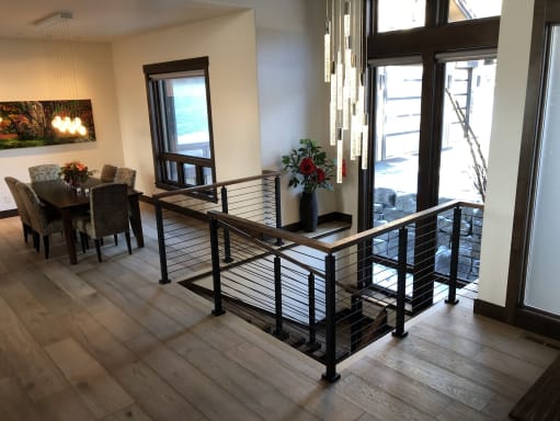 Black rod railing with wood handrail encloses a stair opening between a dining area and entry with tall windows.