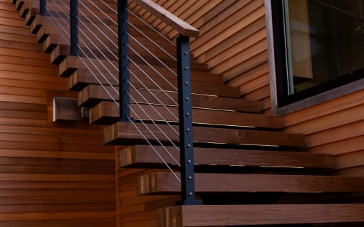 Thick Ipe floating treads with black-post cable railing against warm horizontal wood paneling.