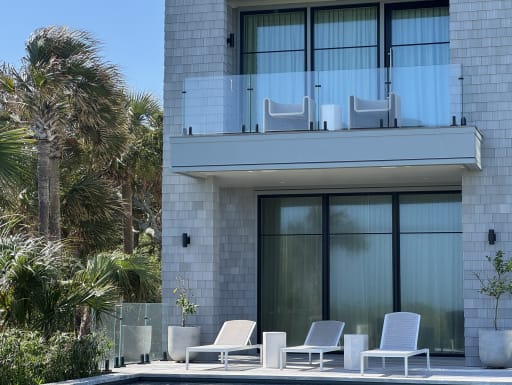 Frameless glass balcony on standoff pins above a poolside patio with lounge chairs and palms