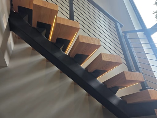 Black mono stringer with warm wood treads lit by LED strips beside black cable railing