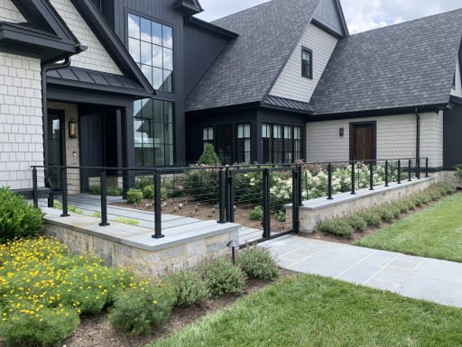 Black cable railing atop a stone wall lines a bluestone garden walk in front of a modern farmhouse