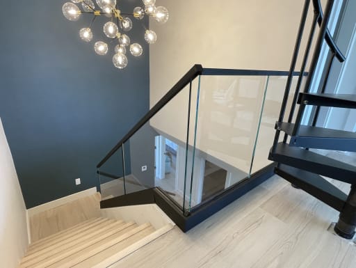 A frameless glass railing with black channel and handrail wraps a stair opening below a globe chandelier and navy wall.