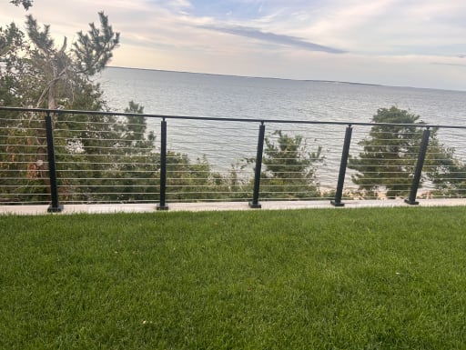 A black cable railing edges a green lawn overlooking a calm bay at dusk.