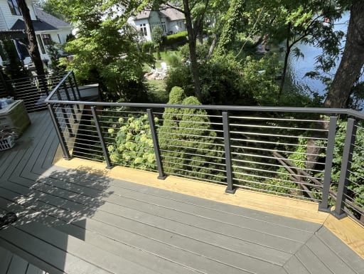 Black cable railing lines the edge of a gray deck overlooking a garden and lake.