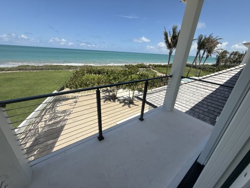 A covered balcony with black cable railing looks out over dunes, palms and the ocean.