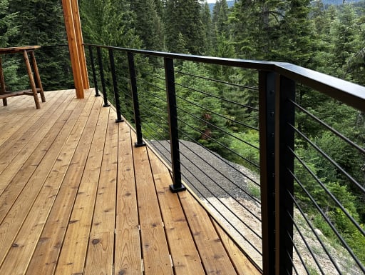 Black cable railing turns a corner on a cedar deck bordered by dense evergreen forest.