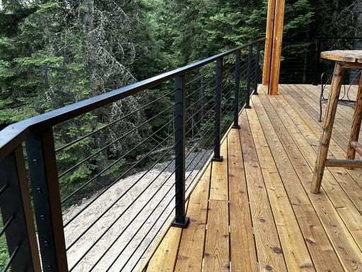 Black cable railing with flat top rail lines a cedar deck beside a rustic table in a forest setting.