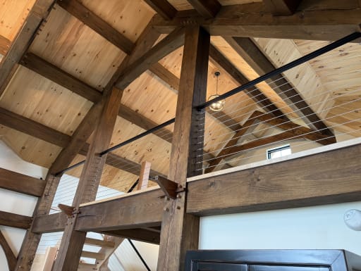 Cable railing with black top rail runs between heavy timber posts in a vaulted pine timber-frame home.