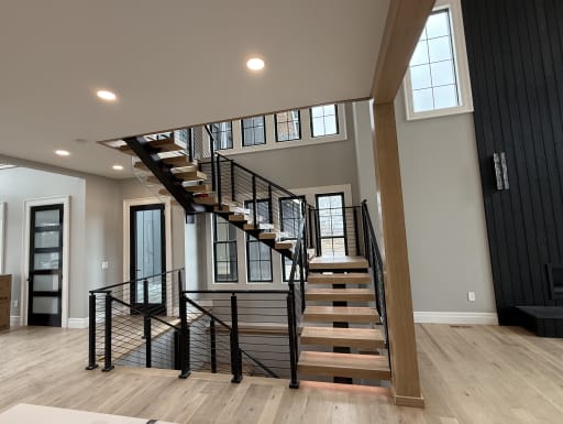 Light oak treads on a black center stringer climb to a landing with black horizontal cable railing beneath tall windows.