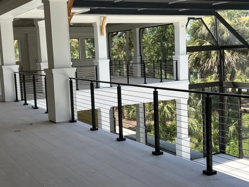 Black posts and top rail with horizontal stainless cables line a wide covered veranda of a white home among palm trees.